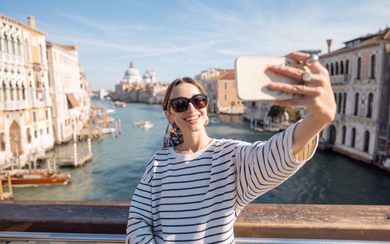 Young woman taking selfie photo on the background of famous Grand Canal in Venice. Idea of spending summer time and happy travel in Italy. Caucasian female in white sunglasses and striped vest